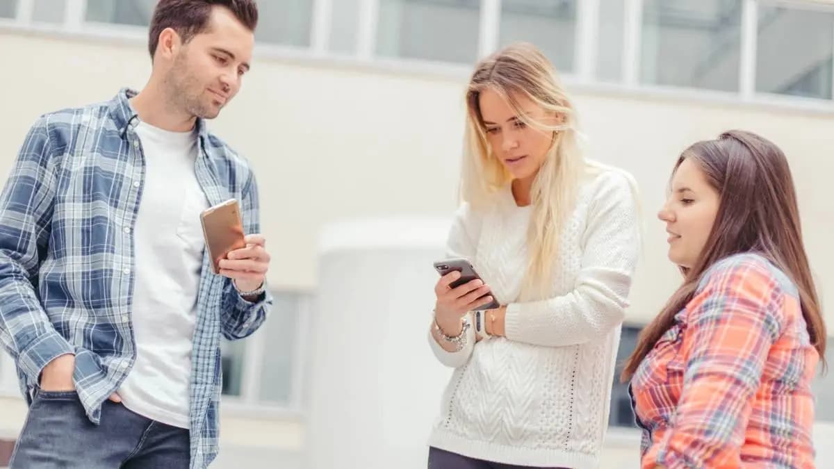 Tři mladí lidé stojí venku a čtou zprávy v mobilech Tři mladí lidé stojí venku a čtou zprávy v mobilech
