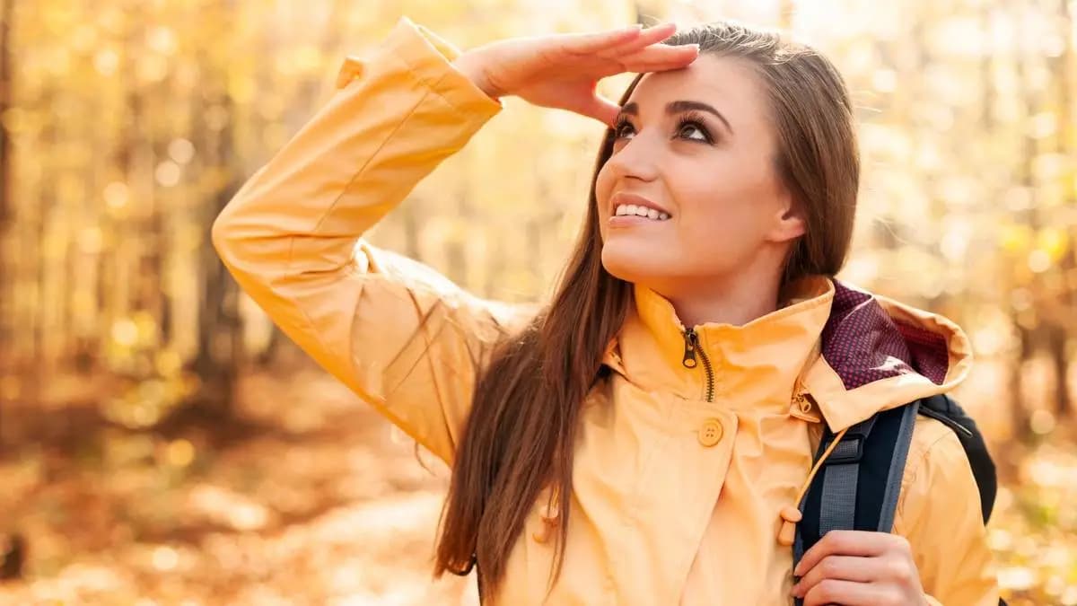 Žena ve žluté bundě s batohem se dívá do slunce v podzimním lese Žena ve žluté bundě s batohem se dívá do slunce v podzimním lese